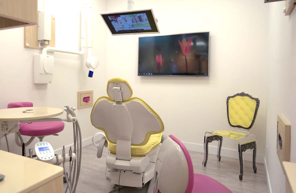 A treatment room at Ironwood Dental Care with a yellow dental chair, pink chairs and an overhead television screen.